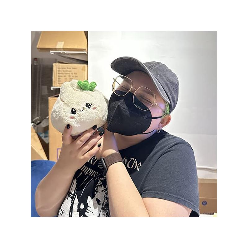 Person with green hair wearing a black mask and glasses smiles while holding a plush Squishable Snugglemi Snackers Soup Dumpling toy.