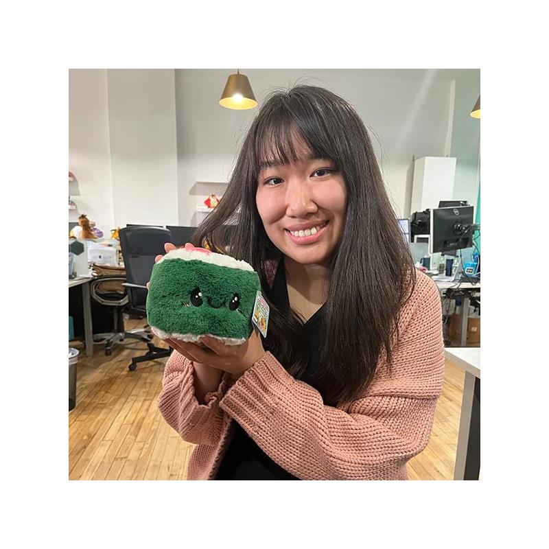 A smiling person holds a plush Squishable Snugglemi Snackers Sushi Roll, featuring green fabric with a white top and cute eyes.