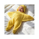 Smiling baby in a hand-crocheted yellow starfish jumpsuit with matching hat, lying on a light-colored bedspread.