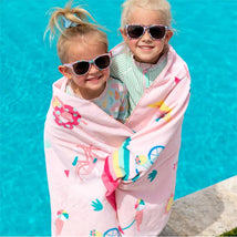 Two young girls, wearing sunglasses, smile while wrapped in a colorful beach towel by a pool.