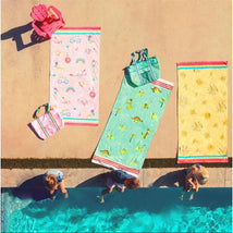 Colorful beach and bath towels with dinosaur and floral designs, along with a matching bag, beside a pool where children play.