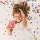 Child in a white dress smiling while holding a pink wallet with a strawberry design, surrounded by confetti.