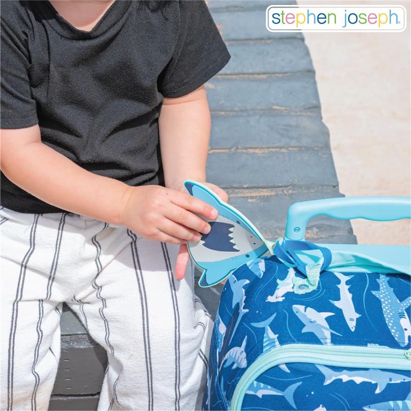 Child attaching a shark-shaped luggage tag to a blue bag covered in shark illustrations.