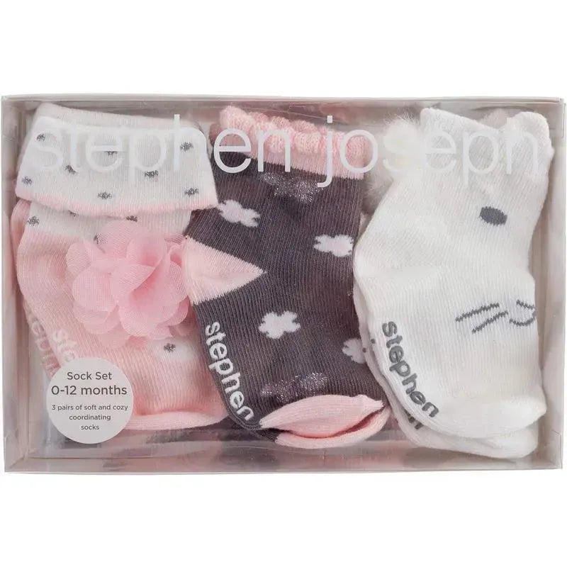 Three pairs of baby socks from Stephen Joseph in a clear box, featuring a pink pair with a flower, a dark pair with clouds, and a white pair with a bunny face.