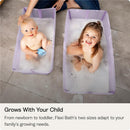 Two young children sit in separate purple bathtubs filled with bubbles. A mother gently observes them.