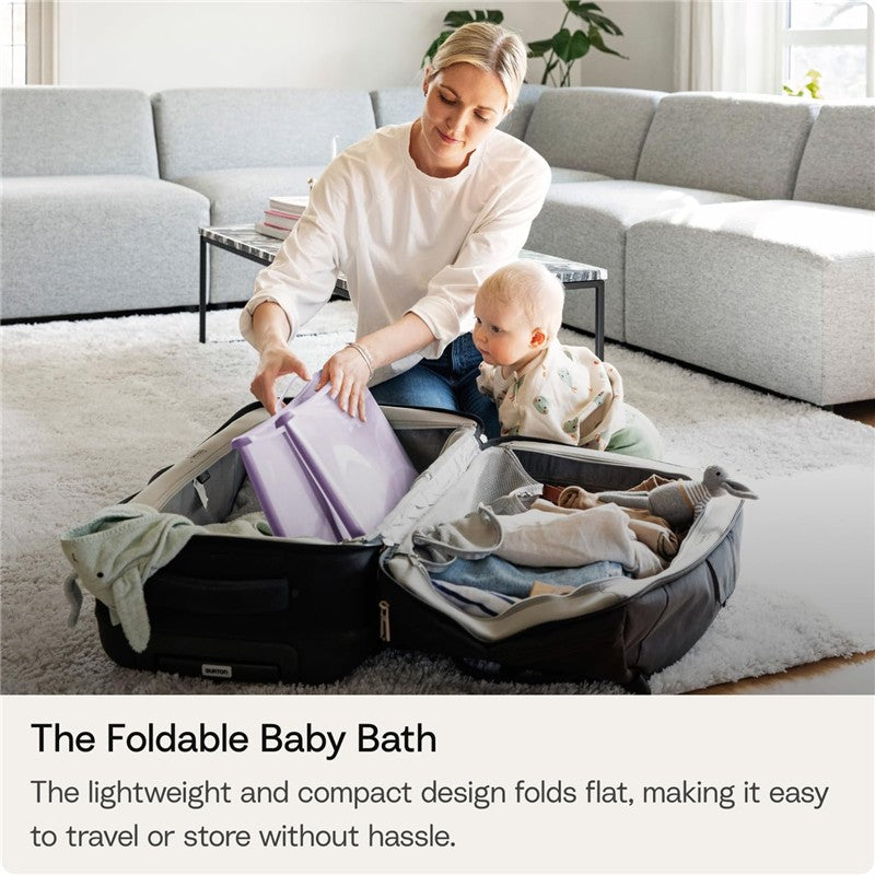 A woman organizes a foldable baby bath in a spacious living room while a baby watches. The bath is purple and compact.