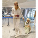 Woman in a white outfit smiles while walking through an airport, holding a passport, as a child in a striped shirt rides on a coral pink Jetkids suitcase.