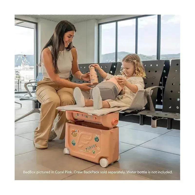 Child seated on Stokke Jetkids™ Bedbox in Moonglow White, while adult assists with a travel item in an airport setting.