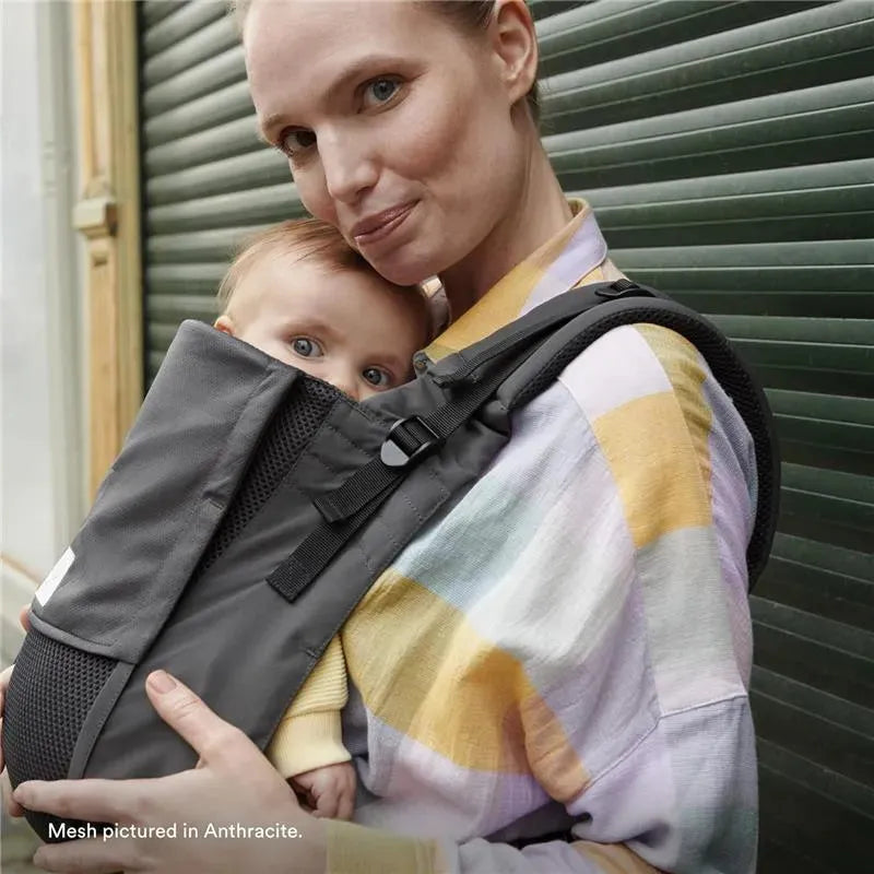 A woman holds a baby in a gray Stokke Limas™ baby carrier, showcasing the mesh design in a city setting.