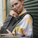 A woman holds a baby in a gray Stokke Limas™ baby carrier, showcasing the mesh design in a city setting.