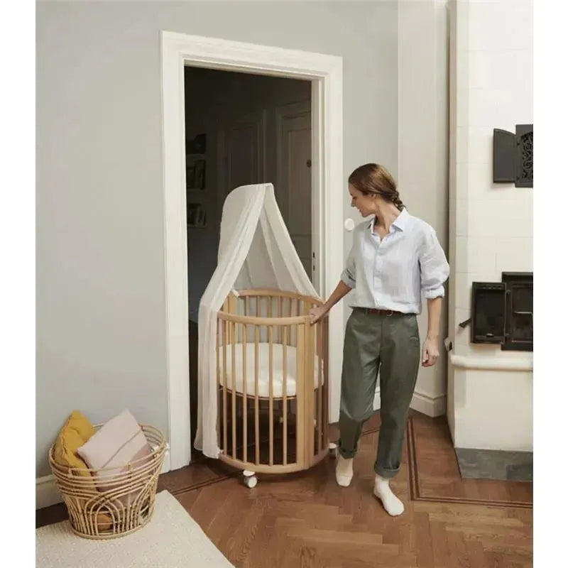 A woman stands next to the Stokke Sleepi Mini Bundle V3 in natural wood, featuring a canopy and a round design, in a cozy room.