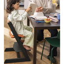 Child sitting in a Stokke Tripp Trapp high chair with an orange cushion, engaged in activities at a table beside an adult.