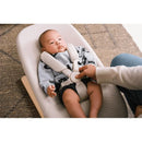 Baby in Stokke Yoga™ Baby Bouncer Light Sand, secured with a harness, while an adult adjusts the safety straps.