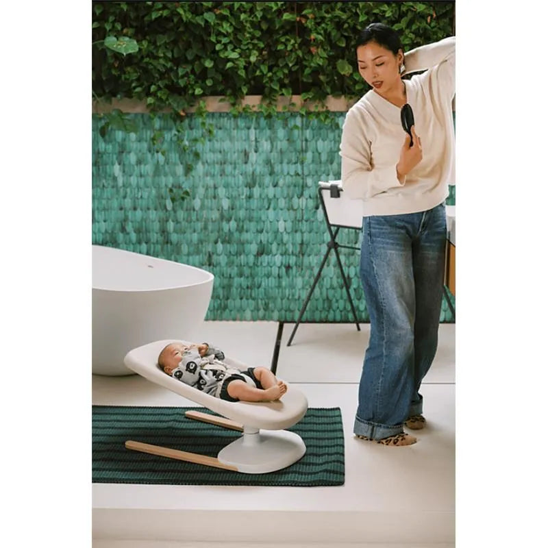 A mother stands beside a Stokke Yoga baby bouncer in light sand, supporting a relaxed baby, with a green textured wall in the background.