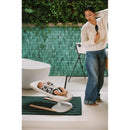 A mother stands beside a Stokke Yoga baby bouncer in light sand, supporting a relaxed baby, with a green textured wall in the background.