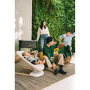 Stokke Yoga™ Baby Bouncer in light sand, with a baby lounging, while a father and child play nearby in a green, cozy setting.