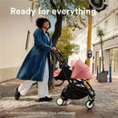 Woman in a denim coat pushing a Stokke Yoyo 0+ Newborn Pack in black with a pink canopy, walking on a city street.