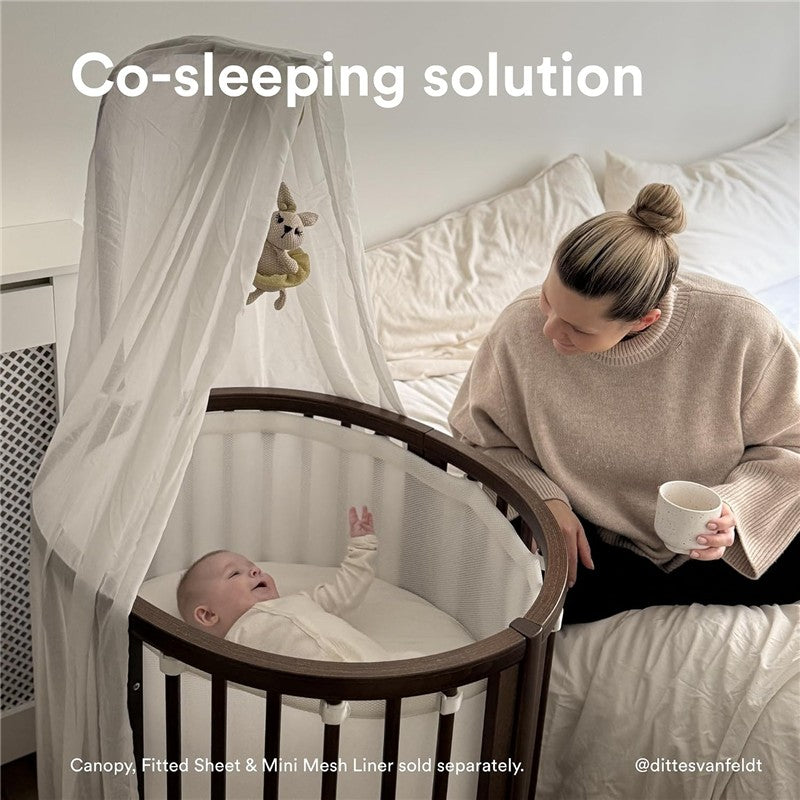 Co-sleeping solution featuring the Stokke® Sleepi™ Mini Bundle in Warm Brown, with a mother watching over a smiling baby in a crib.