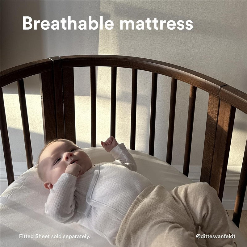 Infant lying in a Stokke® Sleepi™ Mini cradle with a breathable mattress, wearing a white onesie and beige pants.