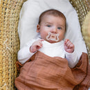 Baby lies in a woven basket, wrapped in a brown blanket, holding a Suavinex silicone pacifier in mouth, looking at the camera.