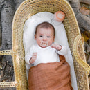 Baby lying in a woven basket, wearing a white outfit and an orange wrap, with a silicone pacifier and a baby bottle nearby.