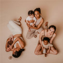 Three women in neutral clothing hold babies in a soft, minimalist setting, emphasizing maternal connection and care.