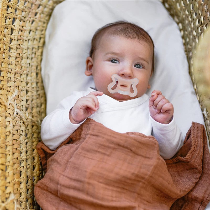 Baby lying in a basket with a brown blanket, holding a transparent silicone pacifier.