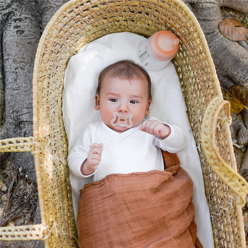 Baby in a woven basket with a brown blanket, holding a Suavinex silicone pacifier and a bottle nearby.