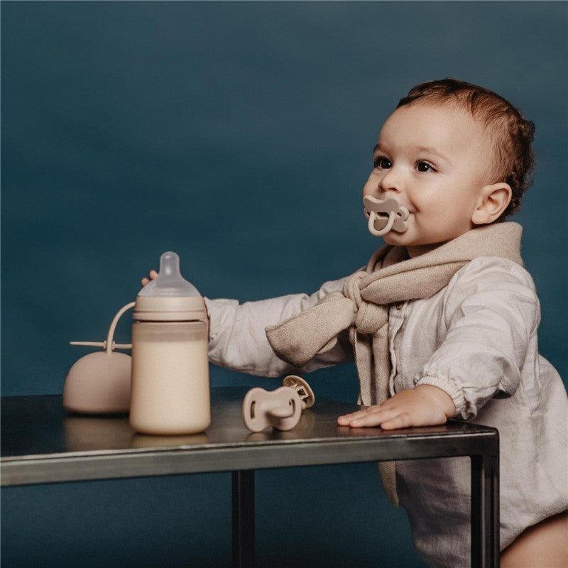 Baby wearing a beige scarf, sitting at a table with a Suavinex Color Essence SX Pro™ Silicone Bottle, pacifiers, and a soft background.