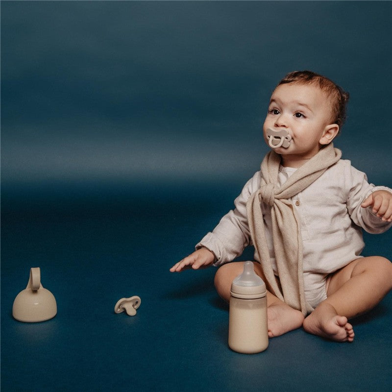 Baby sitting on a blue surface, wearing a light outfit and scarf, with a Suavinex silicone bottle and accessories nearby.