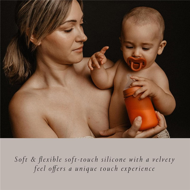 A mother holds a baby with a pacifier, while the baby reaches for a soft orange Suavinex silicone bottle.