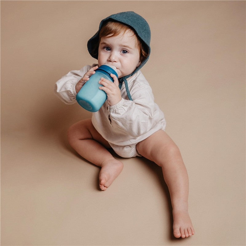 Toddler sitting on a beige background, holding a light blue Suavinex silicone bottle with a blue lid.