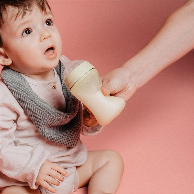 A child sits on a pink background, holding a Suavinex Color Essence SX Pro™ Silicone Bottle in Rose Water, appearing surprised.