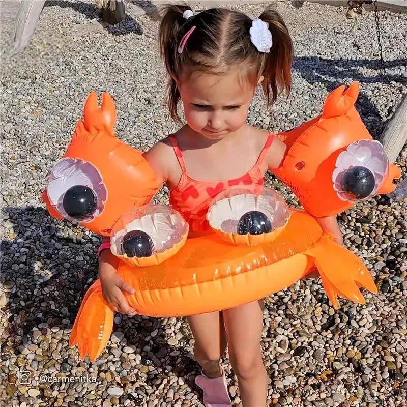Child wearing Sunnylife Buddy Float Bands in the shape of an orange sea creature, standing on a pebbled beach.
