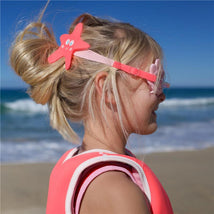Mini swim goggles with a star-shaped strap are worn by a child in a pink swimsuit at the beach, with ocean waves in the background.
