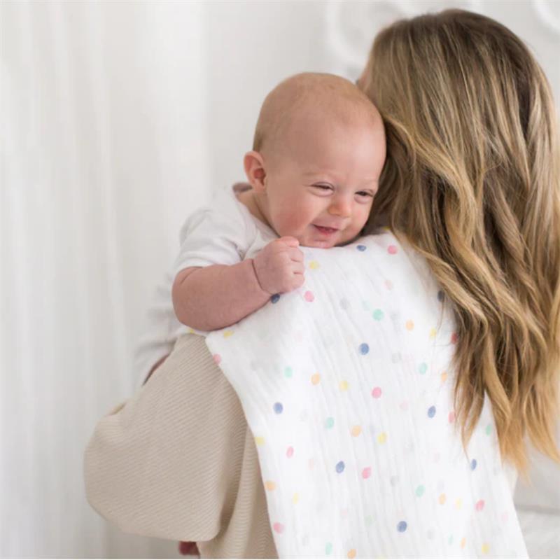 Baby joyfully cuddled in a Swaddle Designs Watercolor Mini Multi Dots muslin swaddle, held by a caregiver.