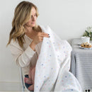 Woman holding a lightweight, colorful polka dot muslin swaddle while seated, with a baby’s feet visible.