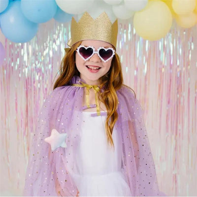 A smiling child wearing heart-shaped sunglasses and a gold crown, dressed in a lavender cape, standing in front of pastel balloons.