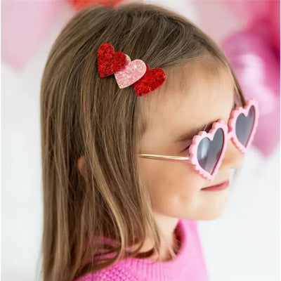A young girl wearing pink heart-shaped sunglasses and a pink sweater, featuring a hair clip with red and pink glitter hearts.