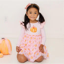 A smiling girl in a pink dress with a pumpkin print, wearing a pumpkin tulle bow headband, sits beside a decorative pumpkin.