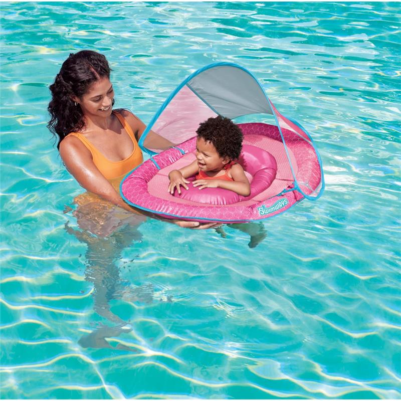 A woman in an orange swimsuit holds a smiling toddler in a pink Swimways baby pool float with a canopy in a swimming pool.