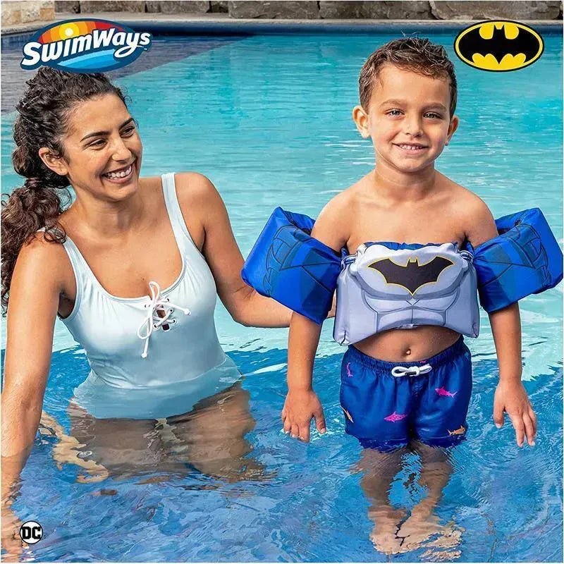A woman in a swimsuit smiles at a young boy wearing a SwimWays Swim Trainer Life Jacket featuring Batman, in a pool.