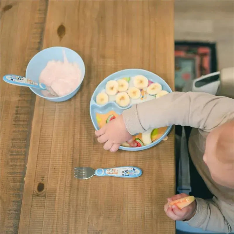 Baby using a Bluey-themed feeding set with banana slices and yogurt on a wooden table.