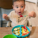 Toddler with short brown hair surprised while holding a fork with a strawberry, sitting at a table with a colorful divided feeding set.