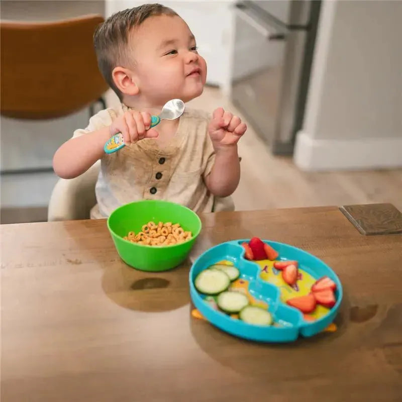 Toddler sitting at a table with a green bowl of cereal and a blue, fish-themed feeding plate with fruits and cucumbers.