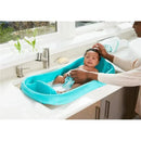 A baby bath with a turquoise color holds a newborn being bathed by an adult, surrounded by a bright kitchen setting.