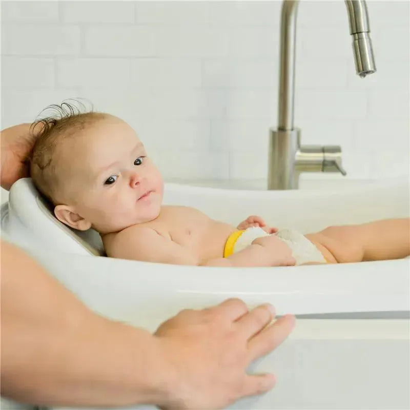Newborn lying in The First Years 3-in-1 baby bathtub, with a hand gently supporting him, in a bright bathroom setting.