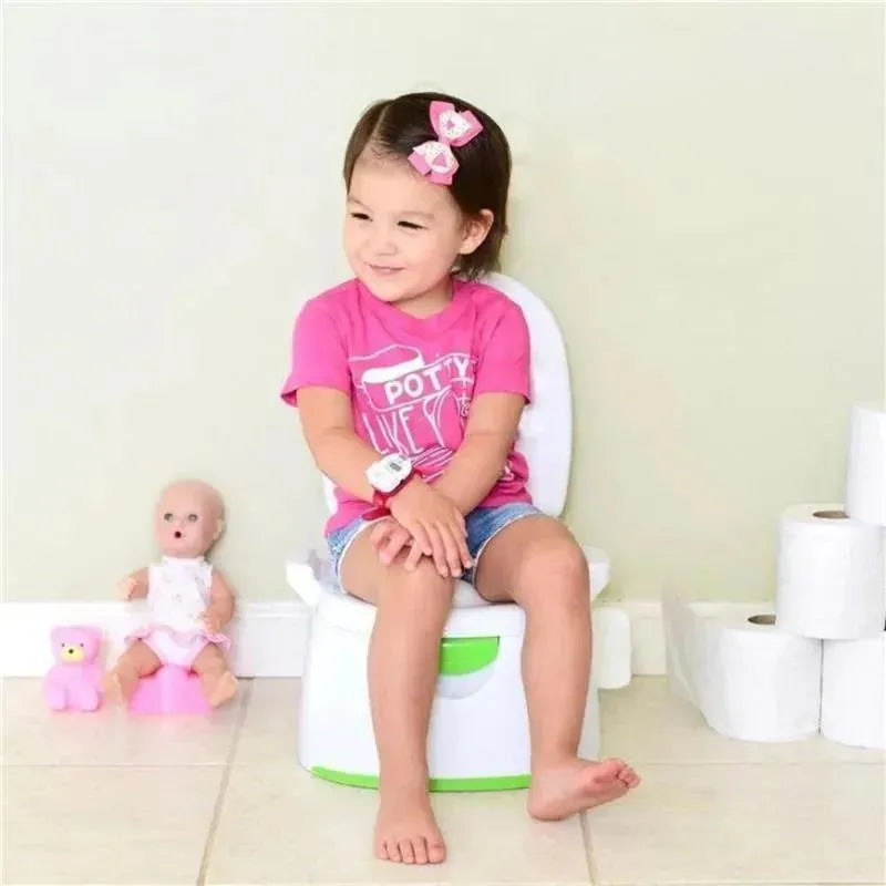 A young girl in a pink shirt sits on a white potty chair, wearing a watch and having a doll and toy bear nearby.