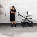 Mother holding a baby, standing next to the Thule Bassinet Mid in blue, set against a neutral wall background.