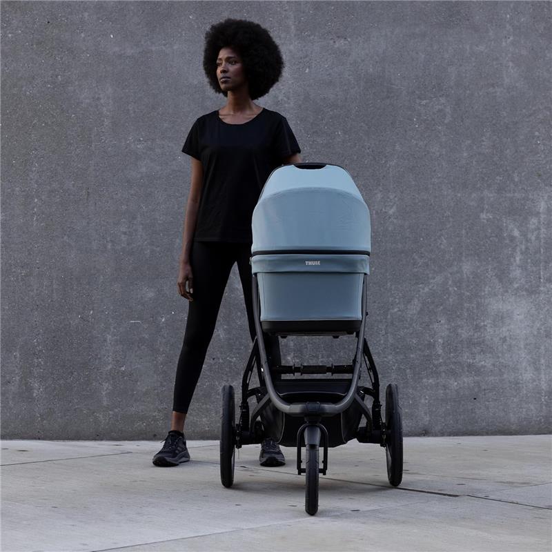 Black woman standing beside Thule Bassinet Mid in blue, featuring a sleek design and sturdy wheels on a concrete background.